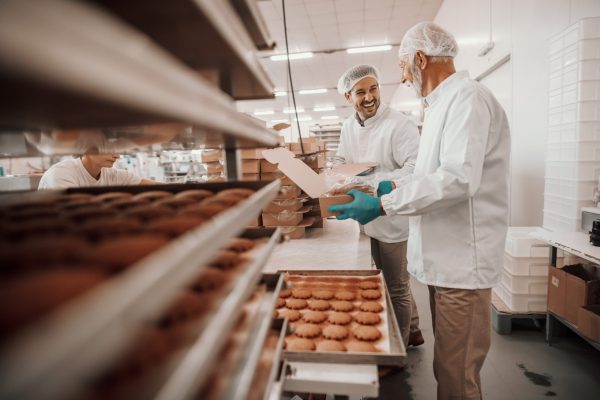 Two hardworking dedicated Caucasian employees dressed in white sterile uniforms collecting and packing cookies in boxes. Food plant interior.